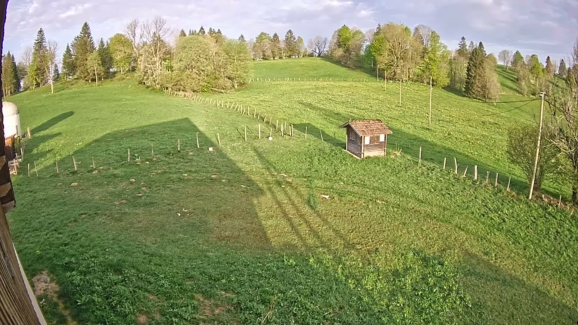 A grassy field with a small shed, a wooden fence, and trees in the background under a partly cloudy sky.