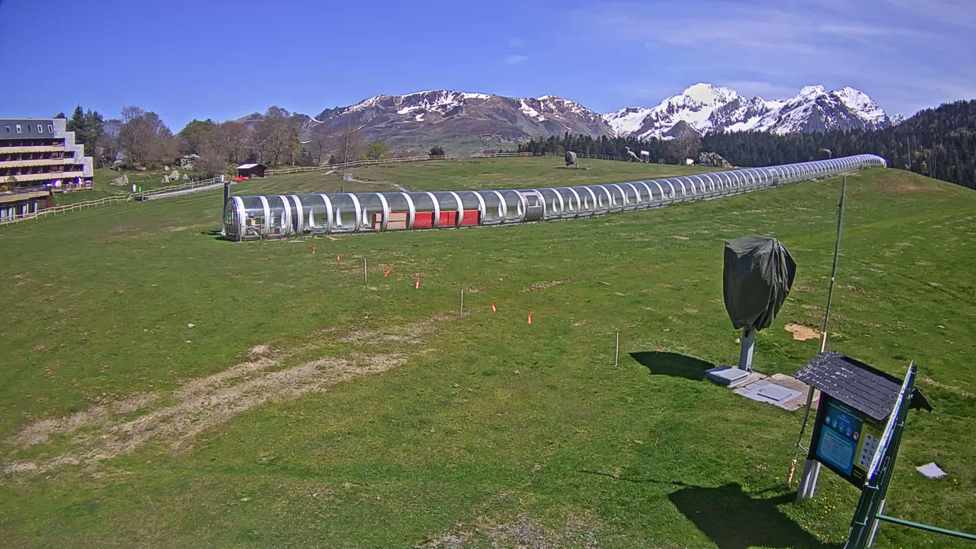 A long, covered structure extends across a grassy field beneath a clear blue sky and snow-capped mountains in the background.
