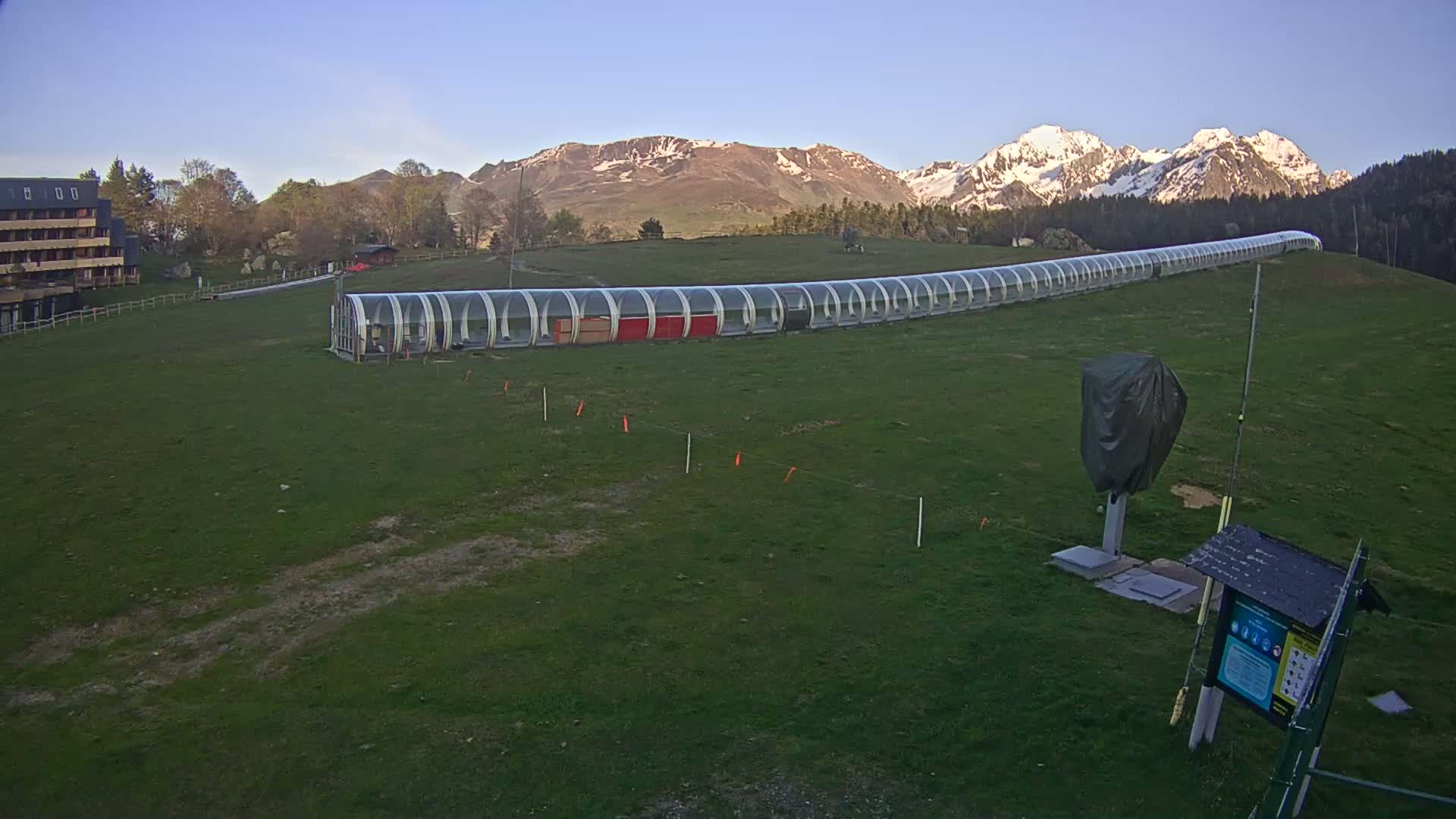 A long, covered walkway extends across a grassy field toward snow-capped mountains under a clear sky.
