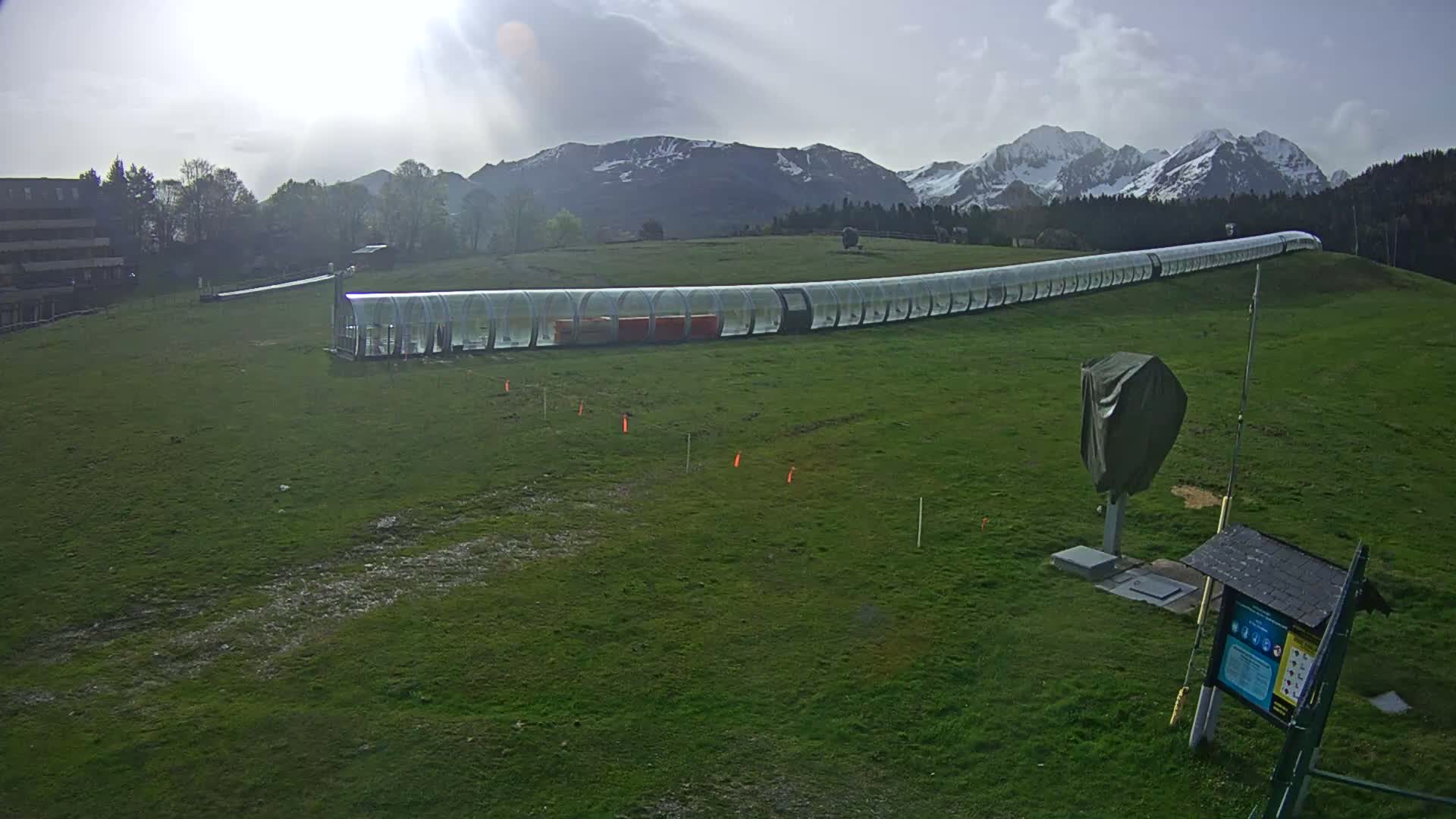 A long, transparent tubular structure sits on a grassy field in front of snow-capped mountains under a partly cloudy sky.