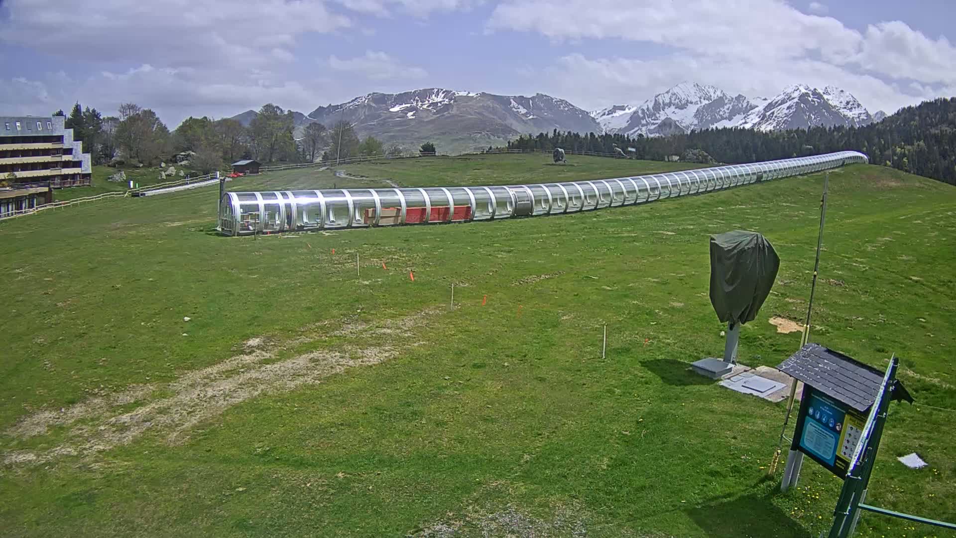 A long, covered conveyor system sits on a grassy hill beneath partly cloudy skies and snow-capped mountains.