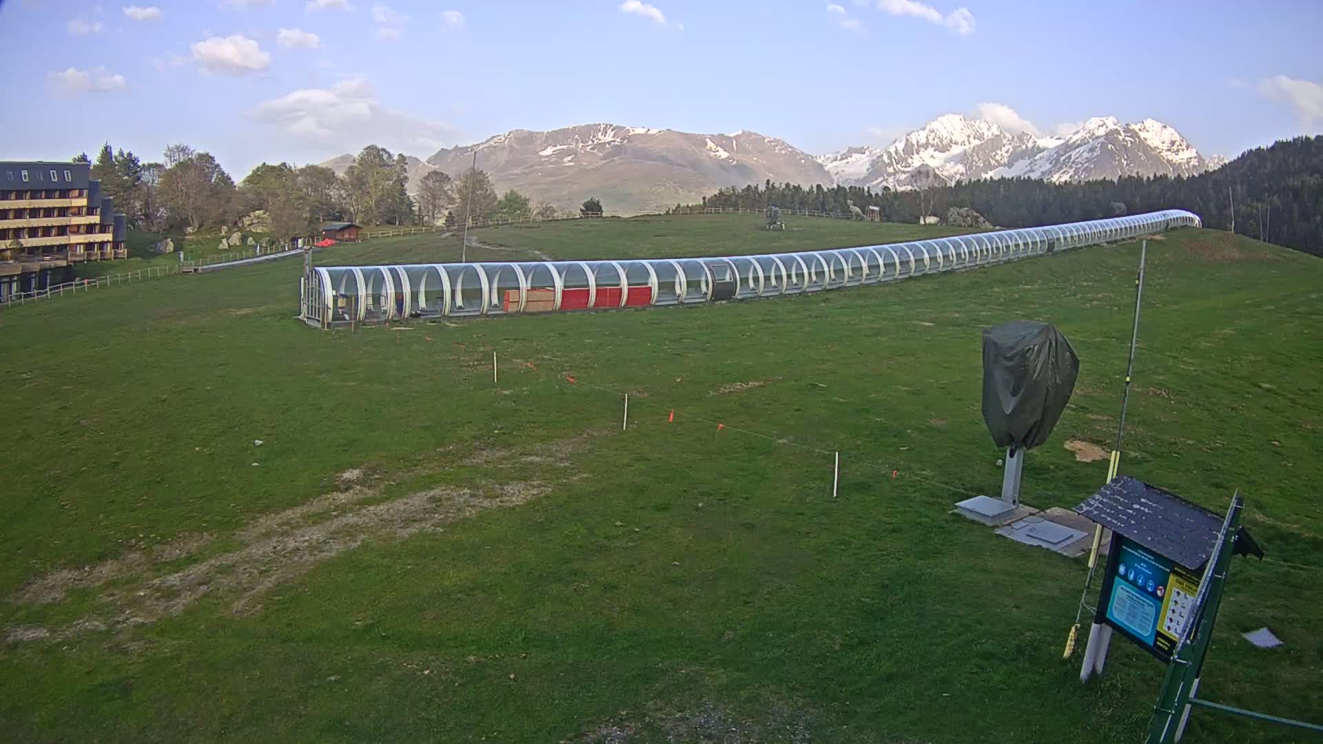 A long, transparent covered walkway stretches across a grassy field, with snow-capped mountains in the background under a partly cloudy sky.