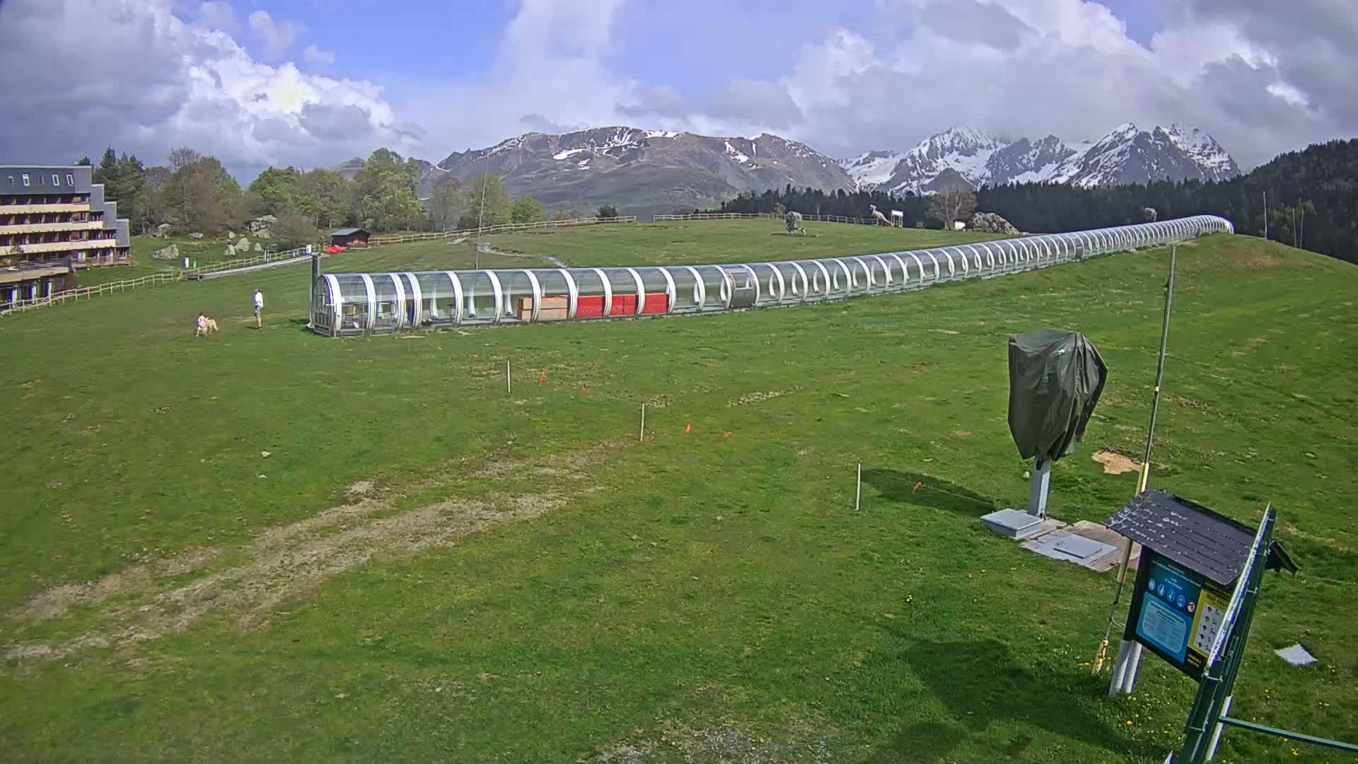 A long, covered transport system extends across a grassy field, with a person and a dog visible nearby, under a partly cloudy sky and snow-capped mountains in the background.