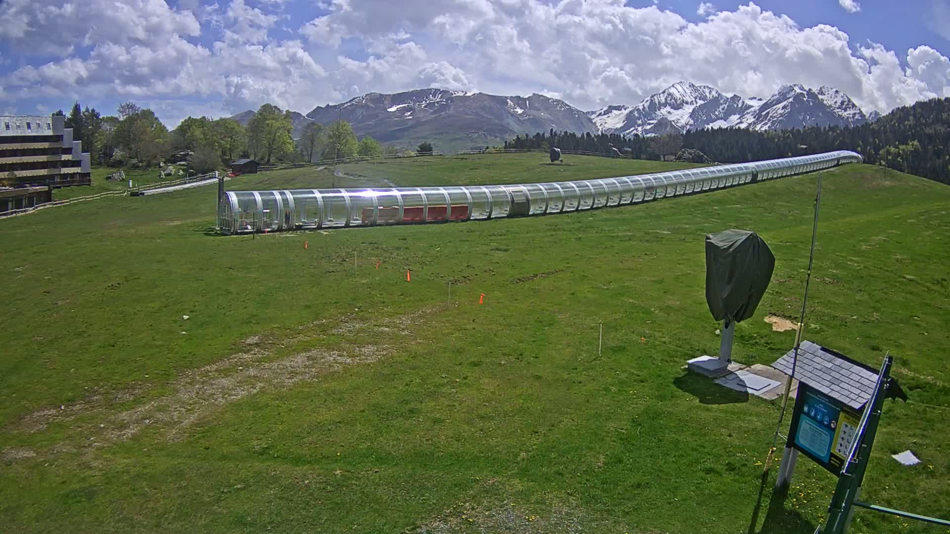 A long, covered lift runs across a grassy field toward snow-capped mountains under a partly cloudy sky.