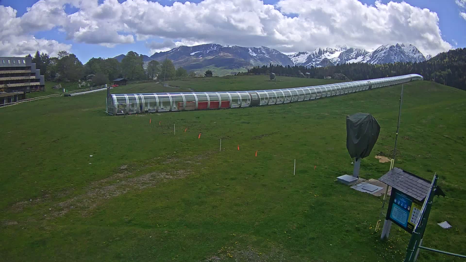 A long, covered moving walkway sits on a grassy hill below snow-capped mountains under a partly cloudy sky.