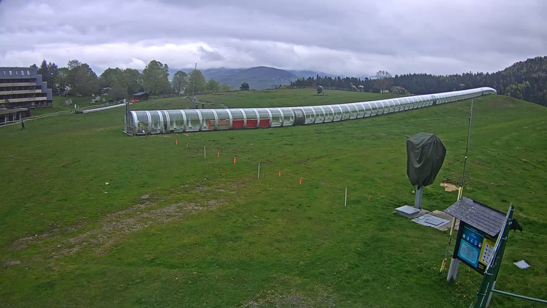 A long, covered, tubular structure sits on a grassy hill under an overcast sky.