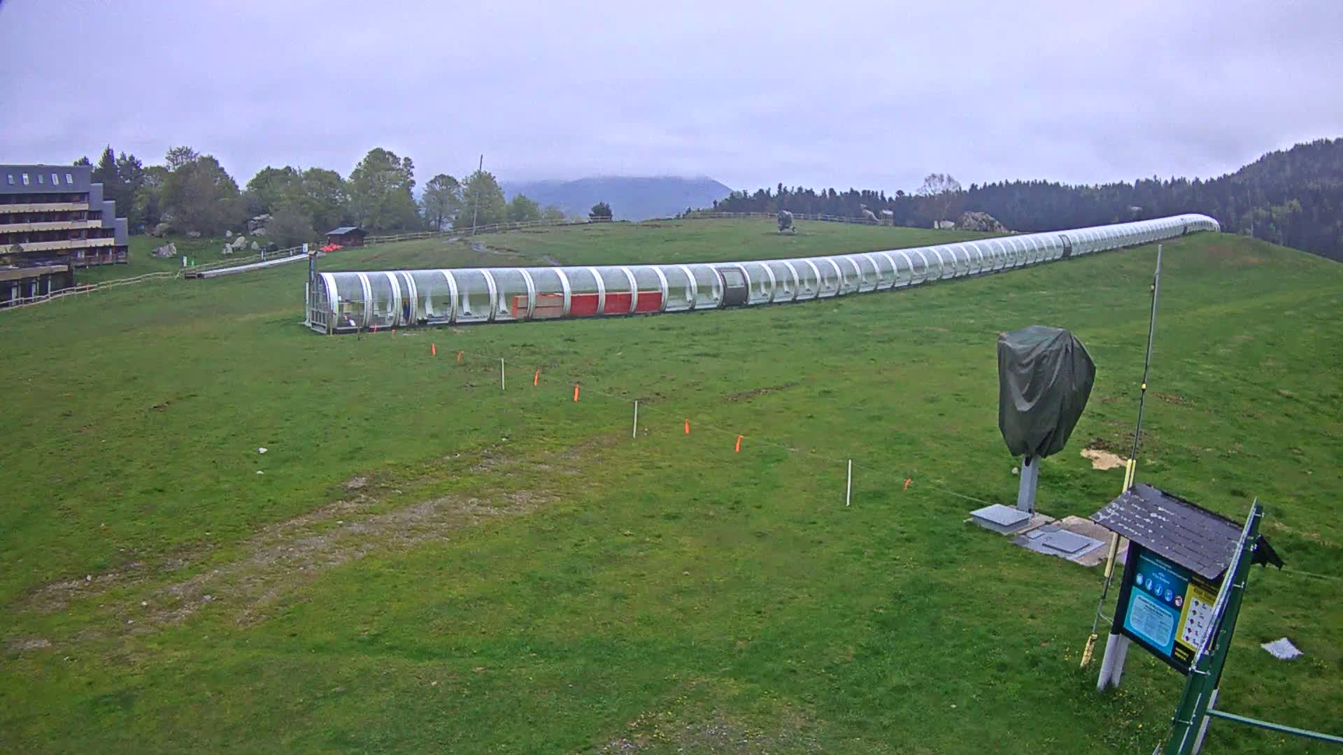 A long, covered conveyance sits on a grassy hill under an overcast sky, with a building visible in the background.