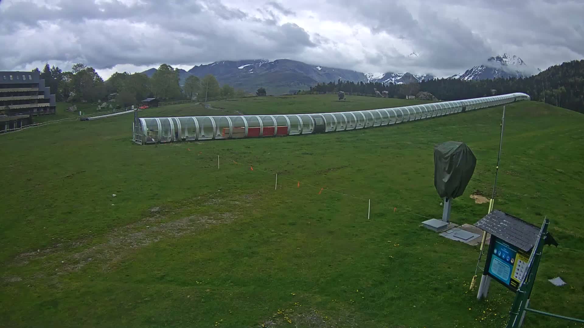 A long, covered conveyor belt-like structure sits on a grassy hill under a cloudy sky, with snow-capped mountains in the background.