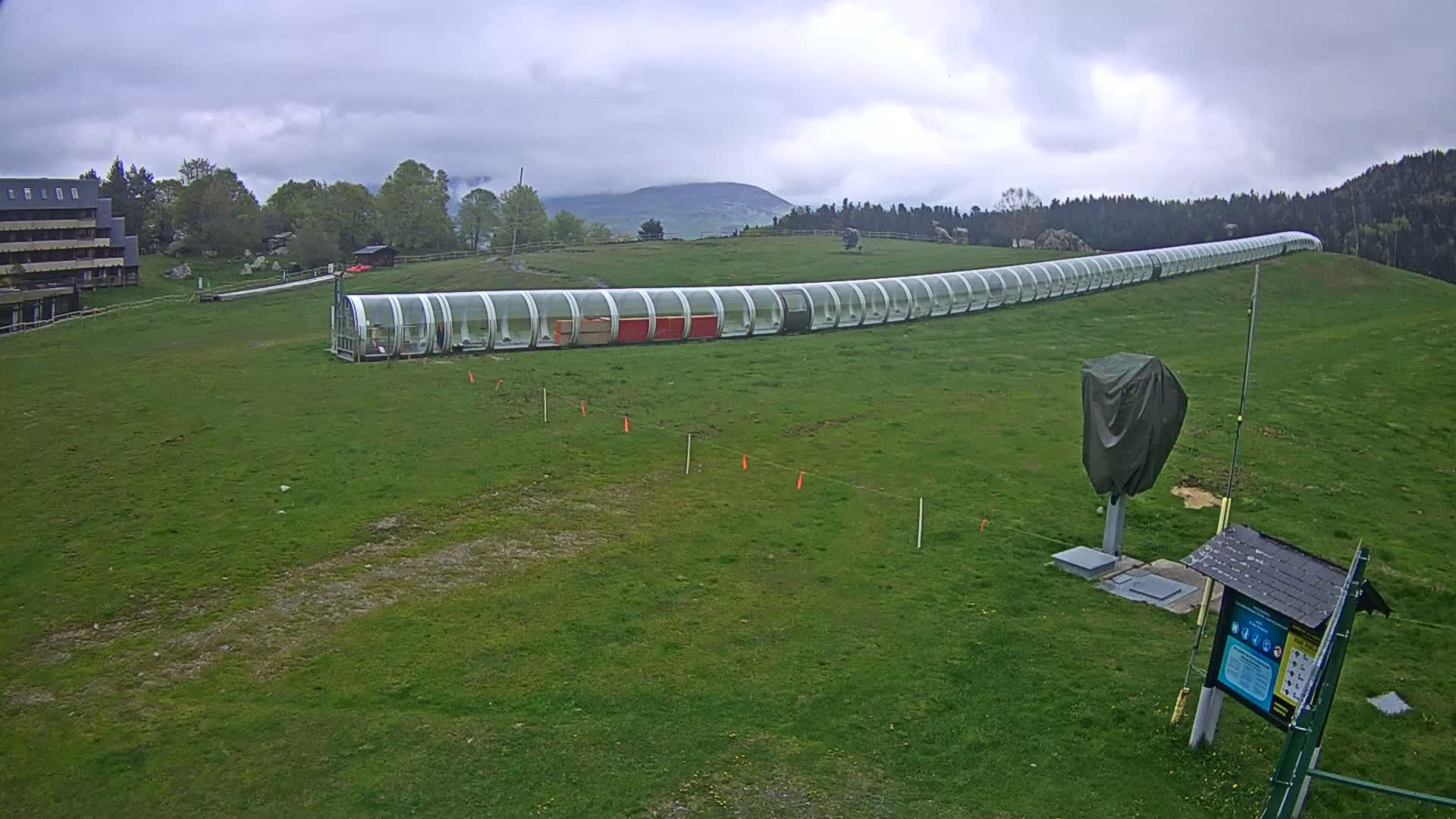 A long, covered walkway stretches across a grassy field under a cloudy sky, with a building and trees visible in the background.