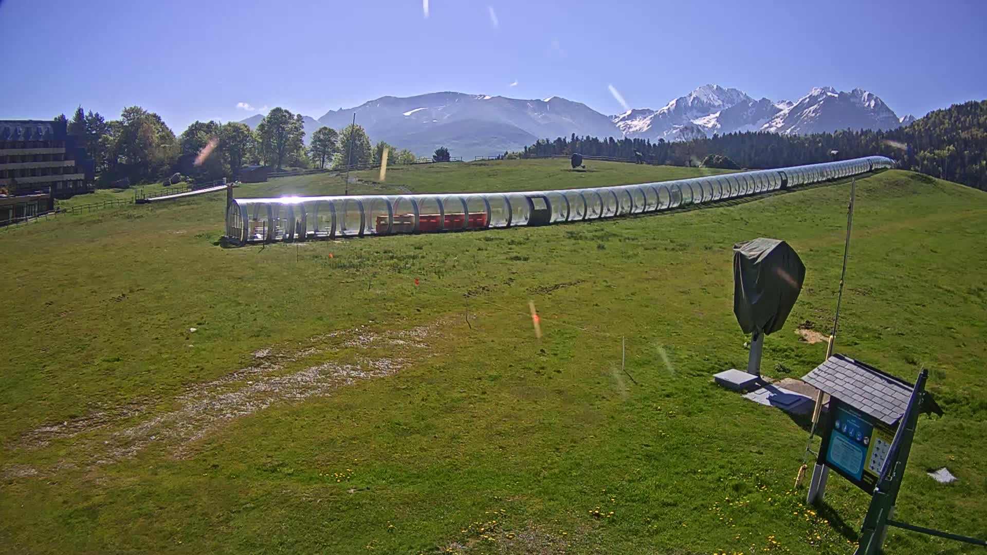 A long, clear tubular structure sits on a grassy hill overlooking snow-capped mountains under a clear blue sky.