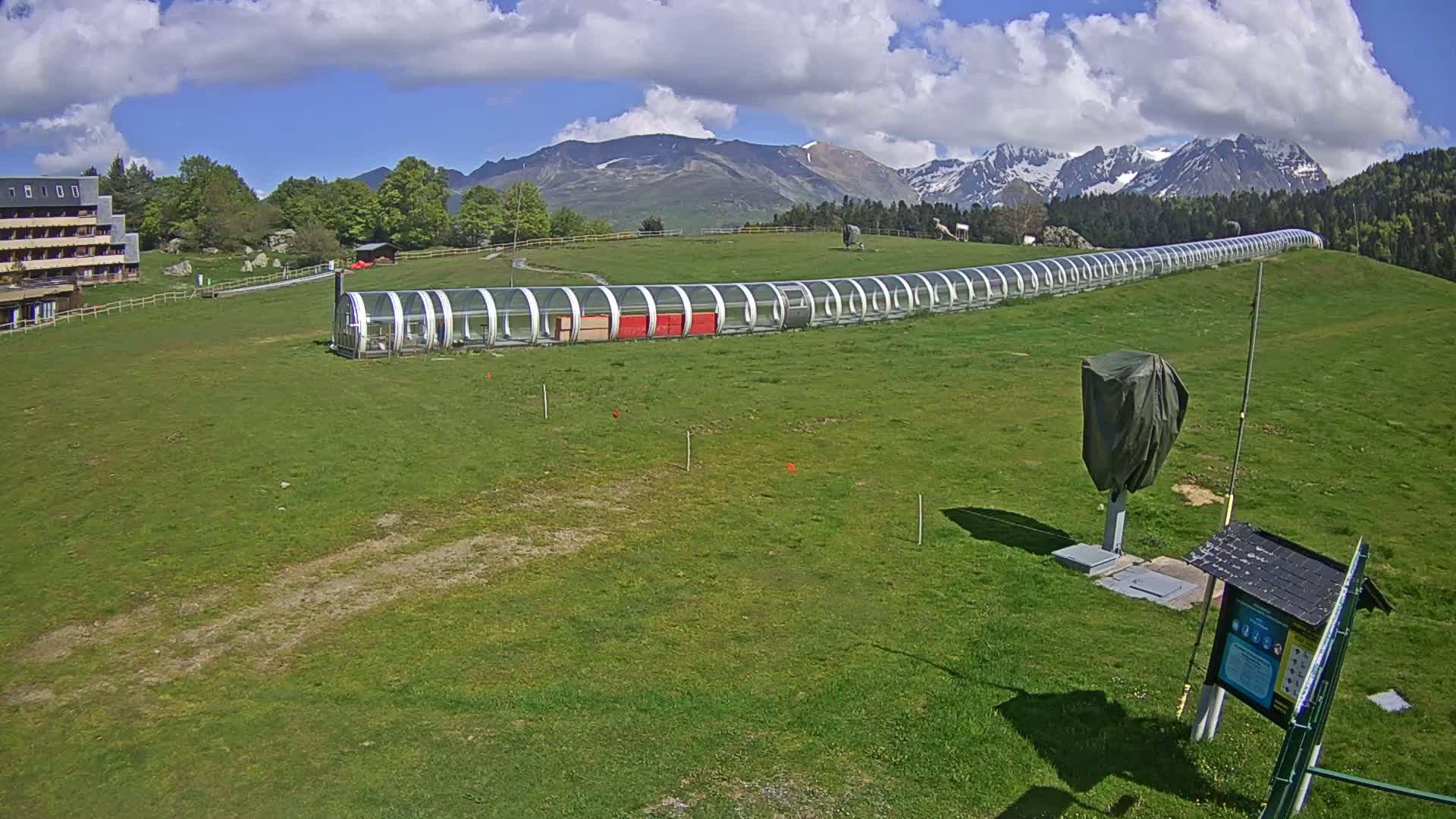 A long, covered structure stretches across a grassy field under a partly cloudy sky, with mountains in the background.