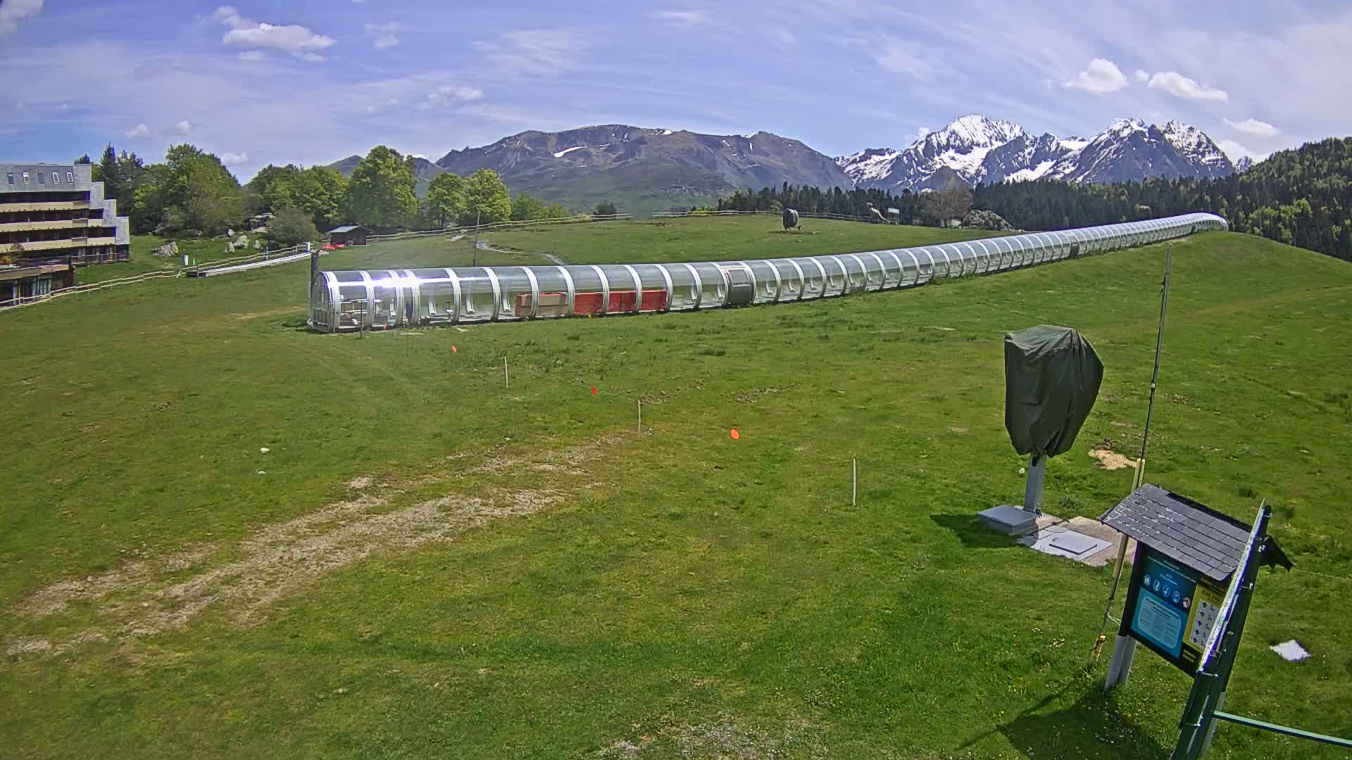 A long, covered transport structure sits on a grassy field in front of snow-capped mountains under a mostly sunny sky.
