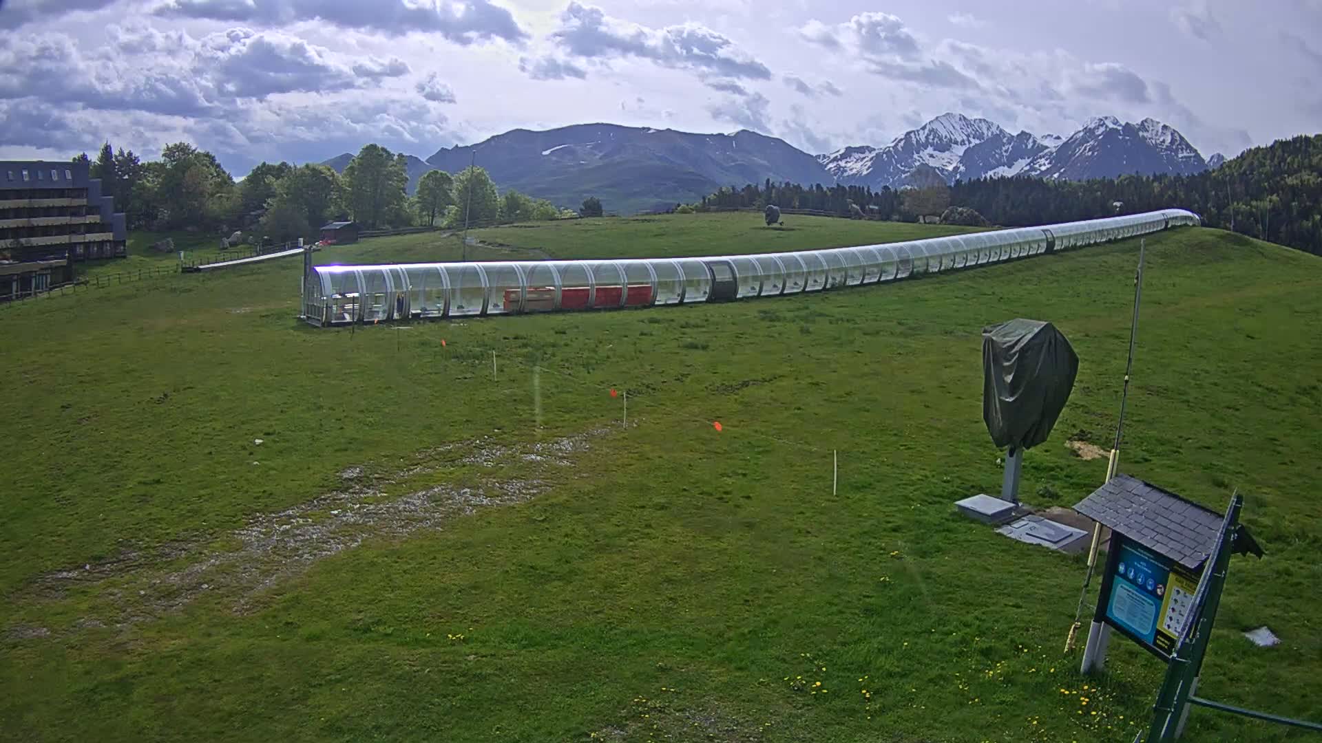 A long, covered conveyor belt system sits on a grassy hill in front of a mountain range under a partly cloudy sky.