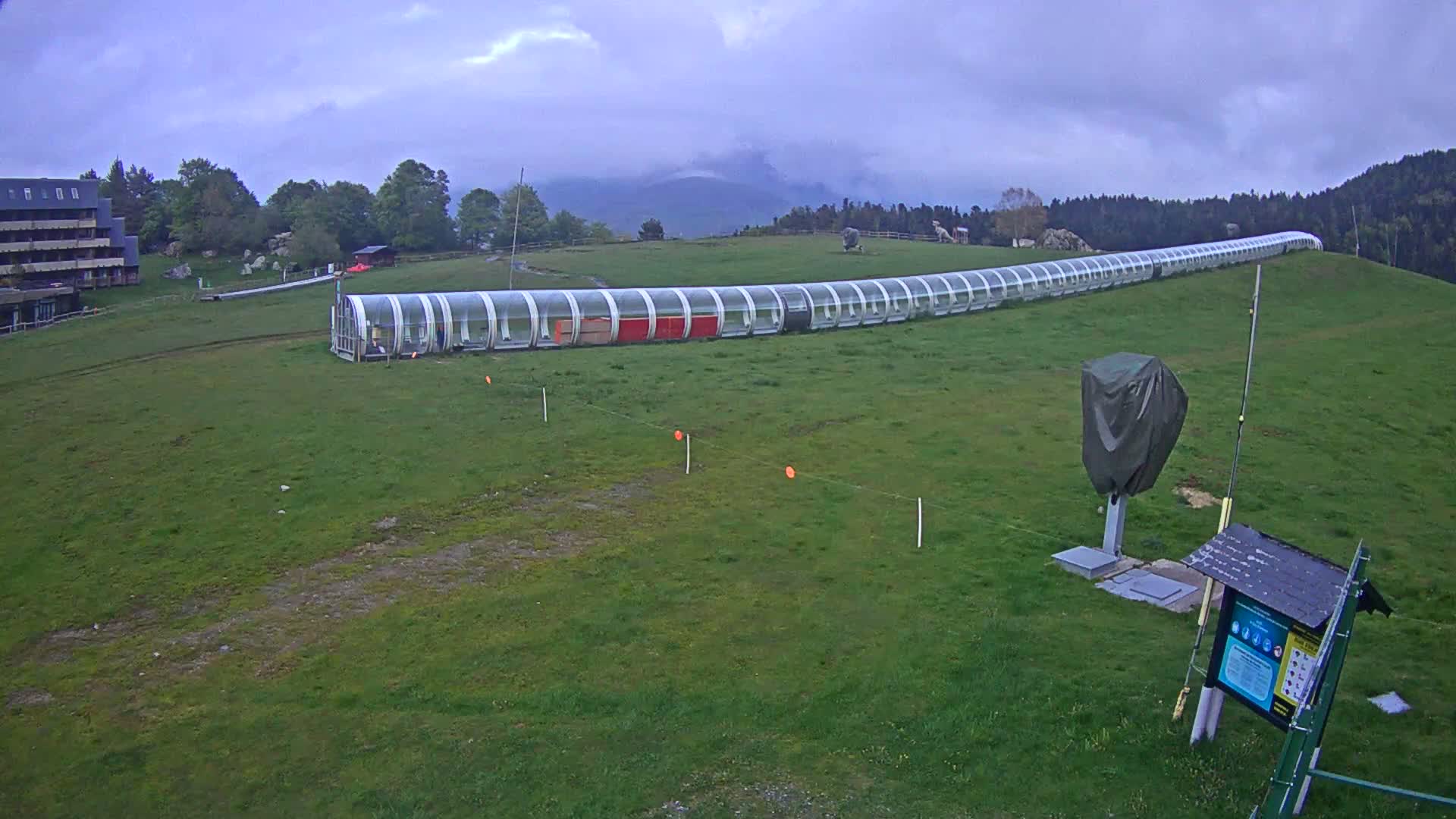 A long, covered conveyor belt-like structure sits on a grassy hill under an overcast sky, near a building and some trees.