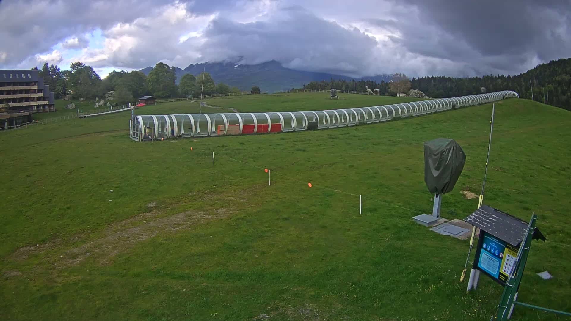 A long, covered toboggan run extends across a grassy field under a cloudy sky, with mountains in the background.