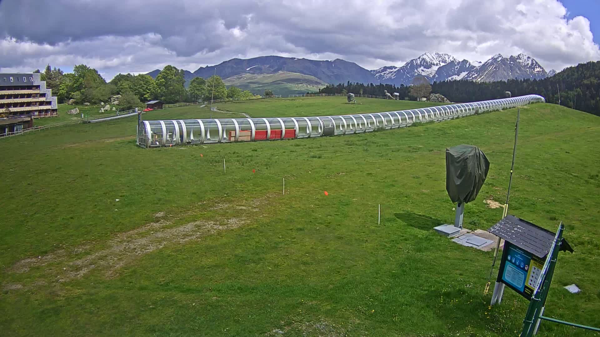 A long, covered transport structure extends across a grassy hillside under a partly cloudy sky, with snow-capped mountains in the background.