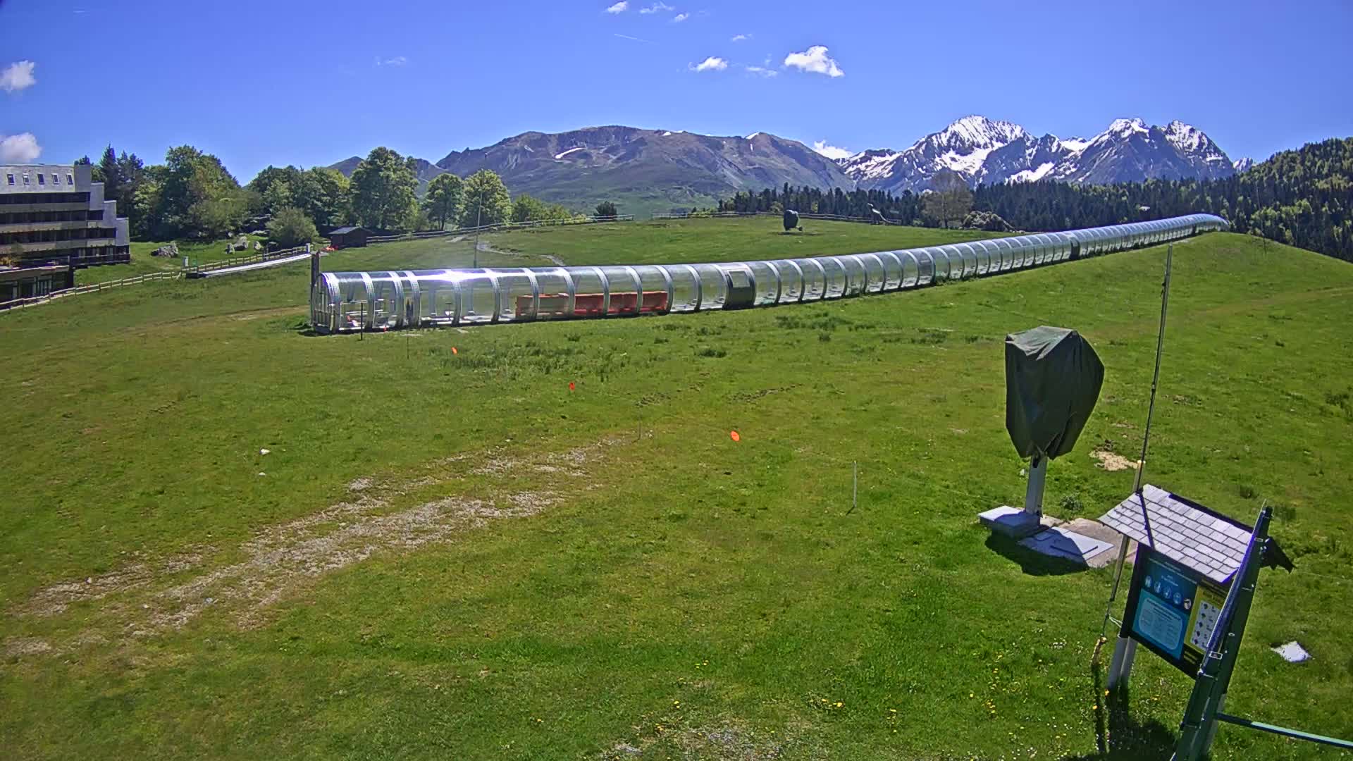 A long, covered transport system is situated on a grassy hillside beneath a clear blue sky and snow-capped mountains.