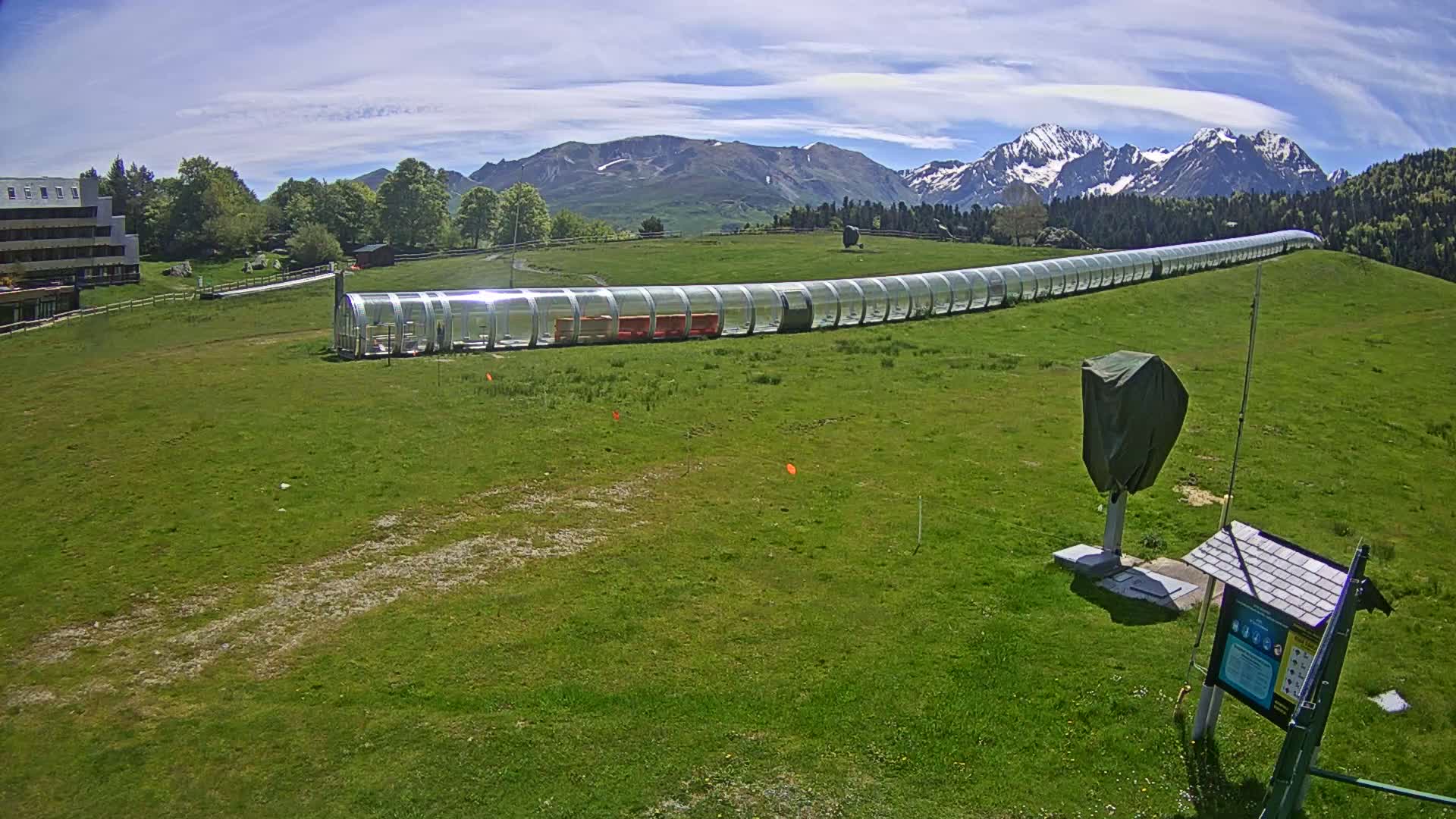 A long, transparent enclosed track sits on a grassy hill overlooking snow-capped mountains under a mostly sunny sky.
