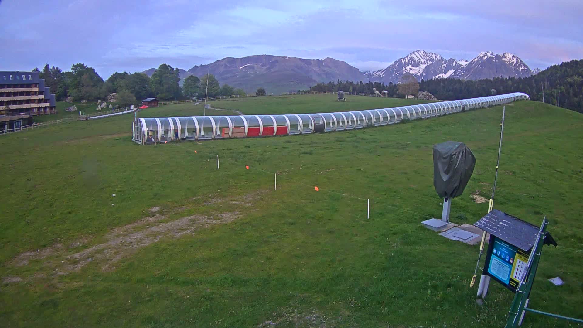A long, covered transport structure extends across a grassy field, with a building and snow-capped mountains visible in the background under a partly cloudy sky.