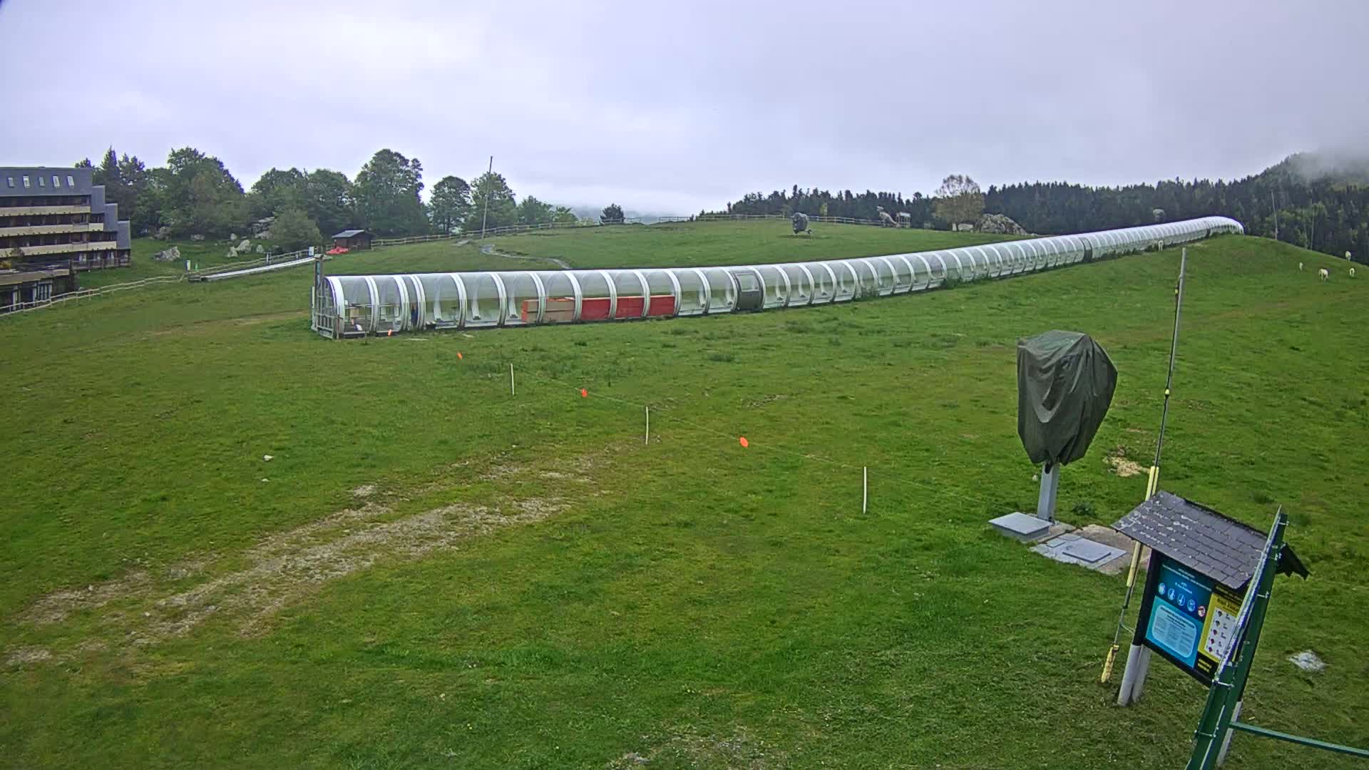A long, covered structure resembling a covered walkway or cable car runs across a grassy hillside under an overcast sky.