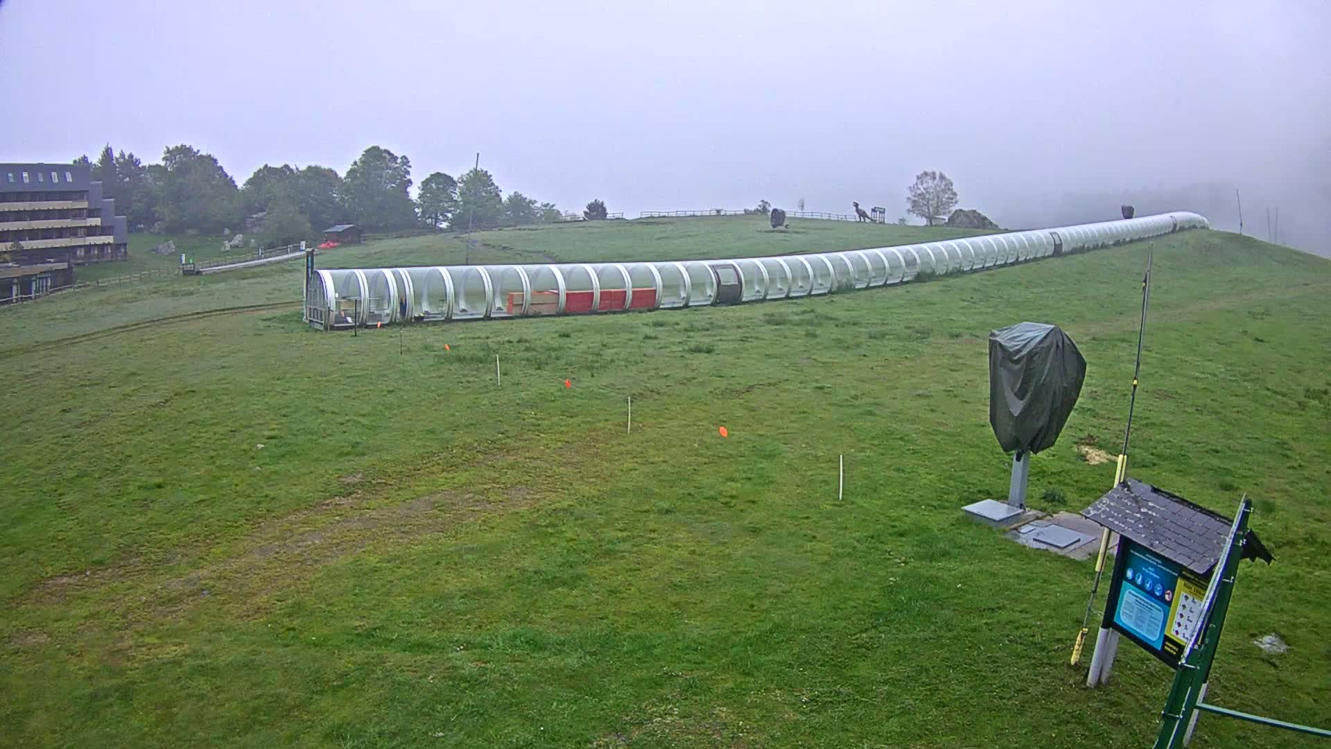A long, covered, tubular structure extends across a grassy hillside in foggy conditions.