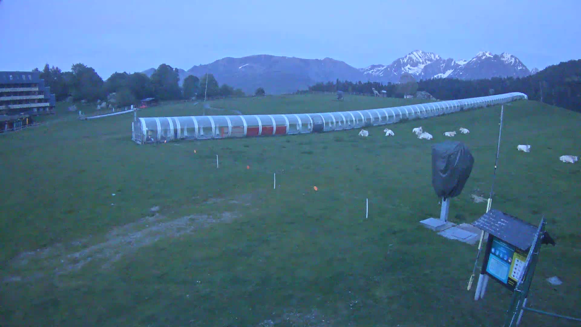 A long, covered structure sits on a grassy field under a twilight sky, with snow-capped mountains in the background.