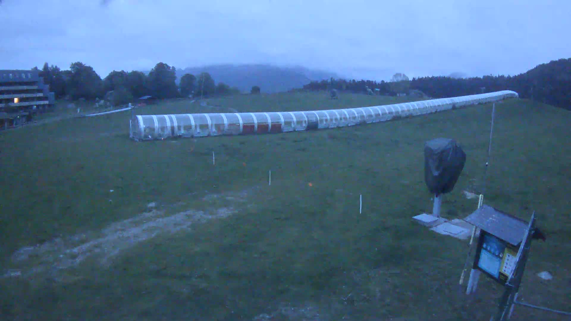 A long, covered structure stretches across a grassy field under an overcast sky.