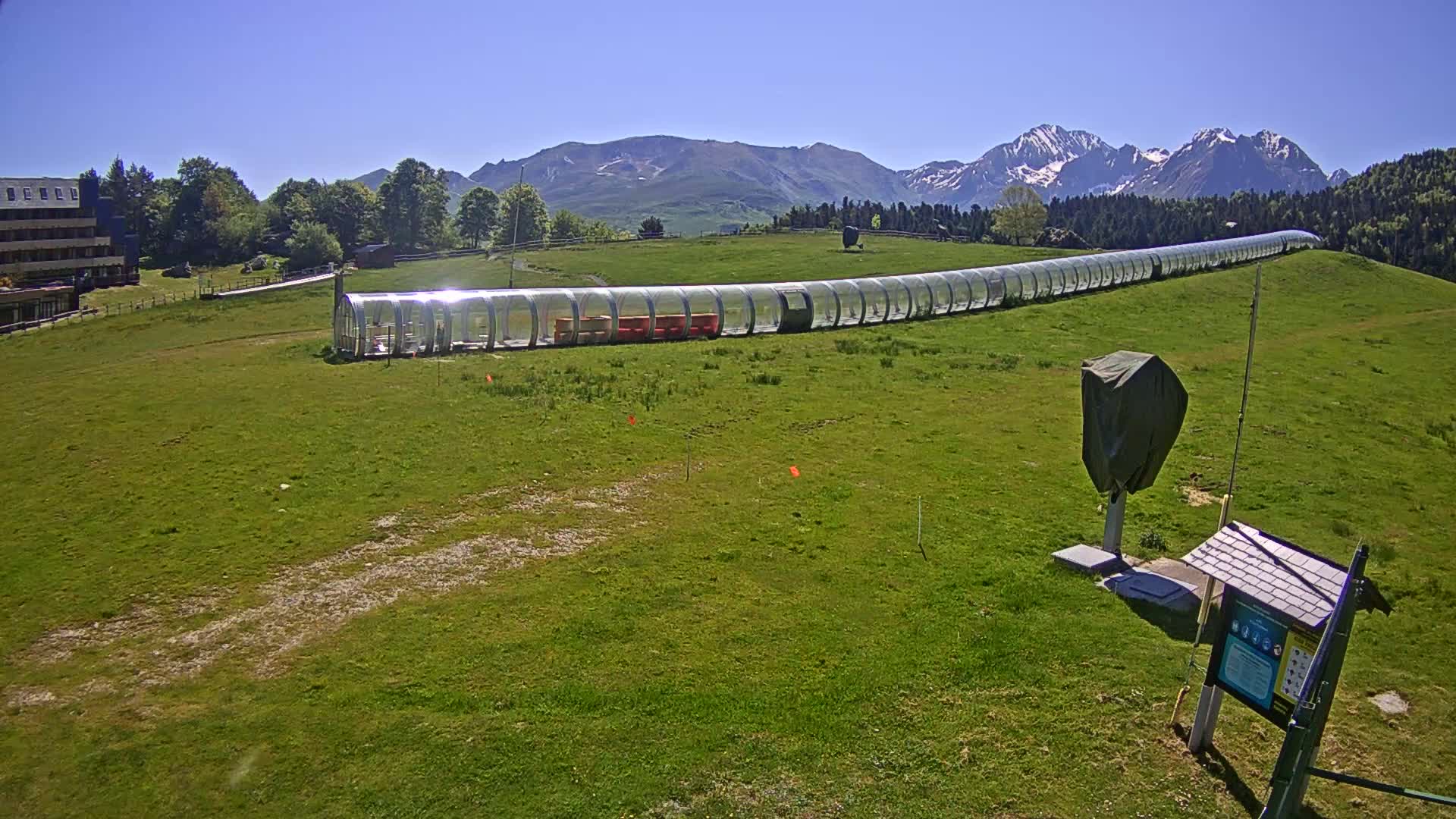 A long, transparent enclosed structure sits on a grassy hill beneath a bright, sunny sky and snow-capped mountains in the distance.