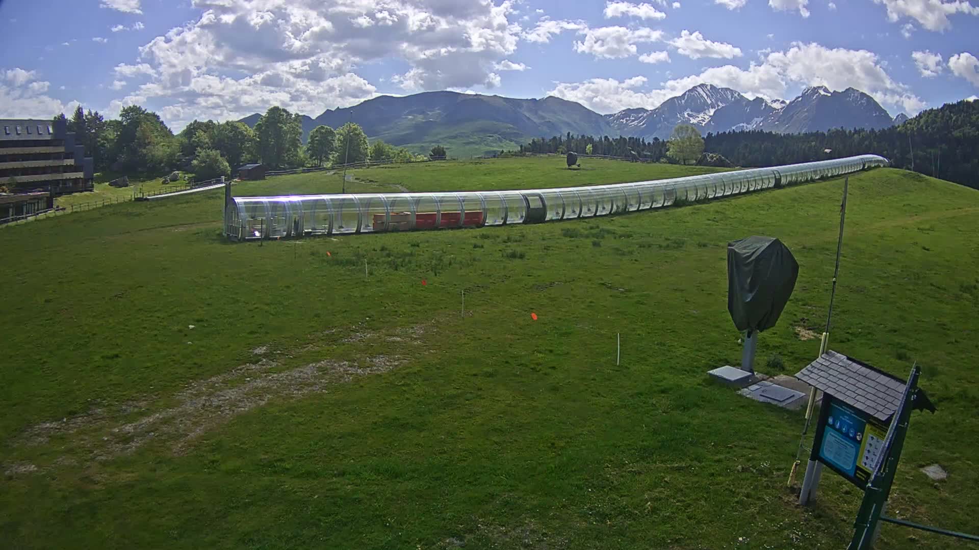 A long, transparent enclosure sits on a grassy hill against a backdrop of mountains under a partly cloudy sky.
