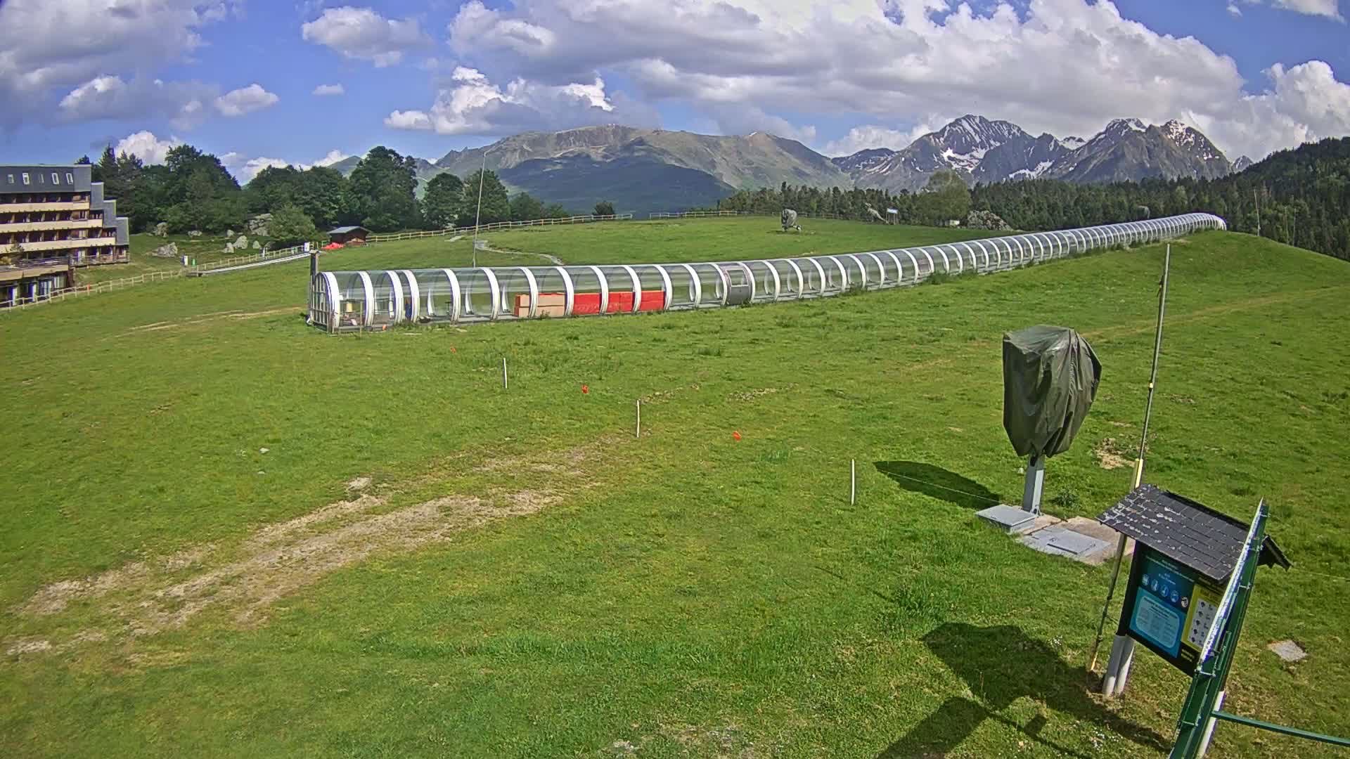 A long, covered structure sits on a grassy field in front of a building and a mountain range under a partly cloudy sky.