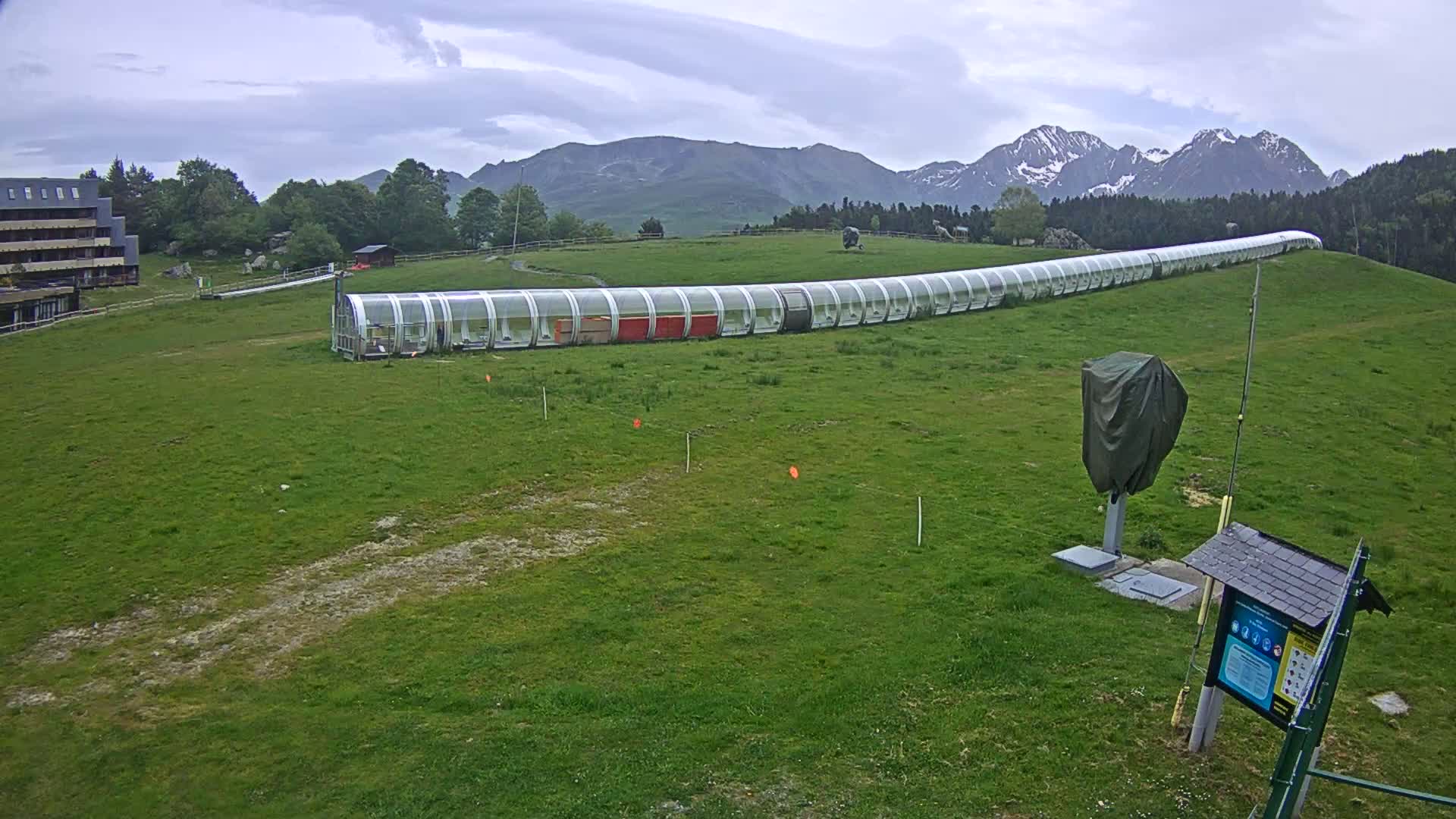 A long, enclosed structure sits on a grassy hill overlooking mountains under a cloudy sky.
