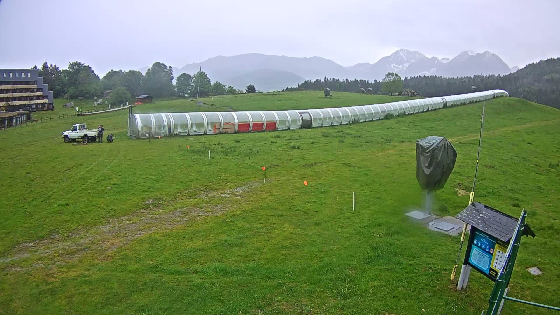 A long, covered structure extends across a grassy field under an overcast sky, with mountains in the background; a truck and a few people are near the structure.
