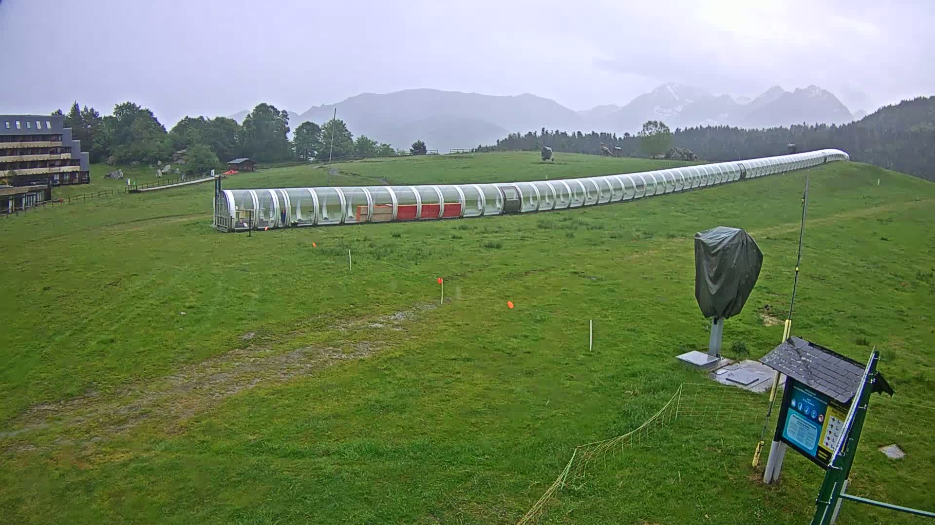 A long, transparent enclosed structure sits on a grassy hillside under an overcast sky, with mountains in the background.