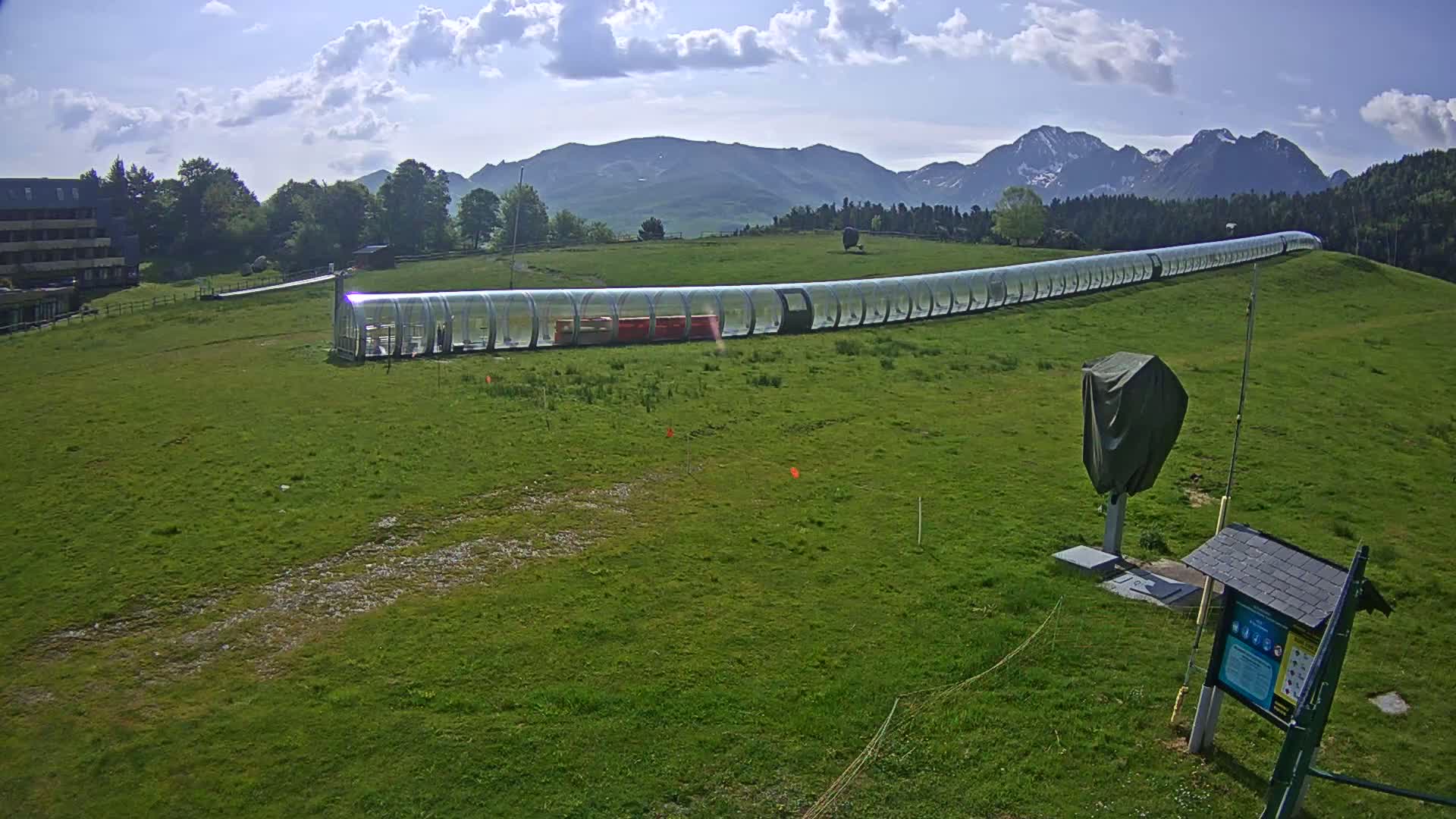 A long, transparent enclosed cable car travels across a grassy field under a partly cloudy sky, with mountains in the background.