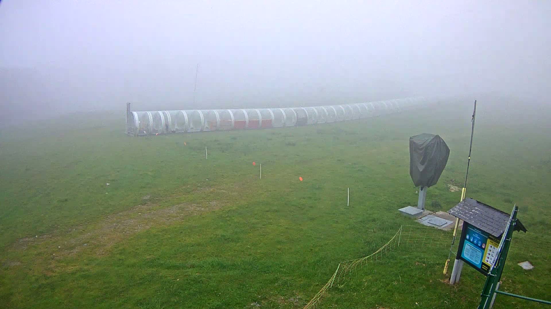 A long, covered structure sits on a grassy field shrouded in thick fog.