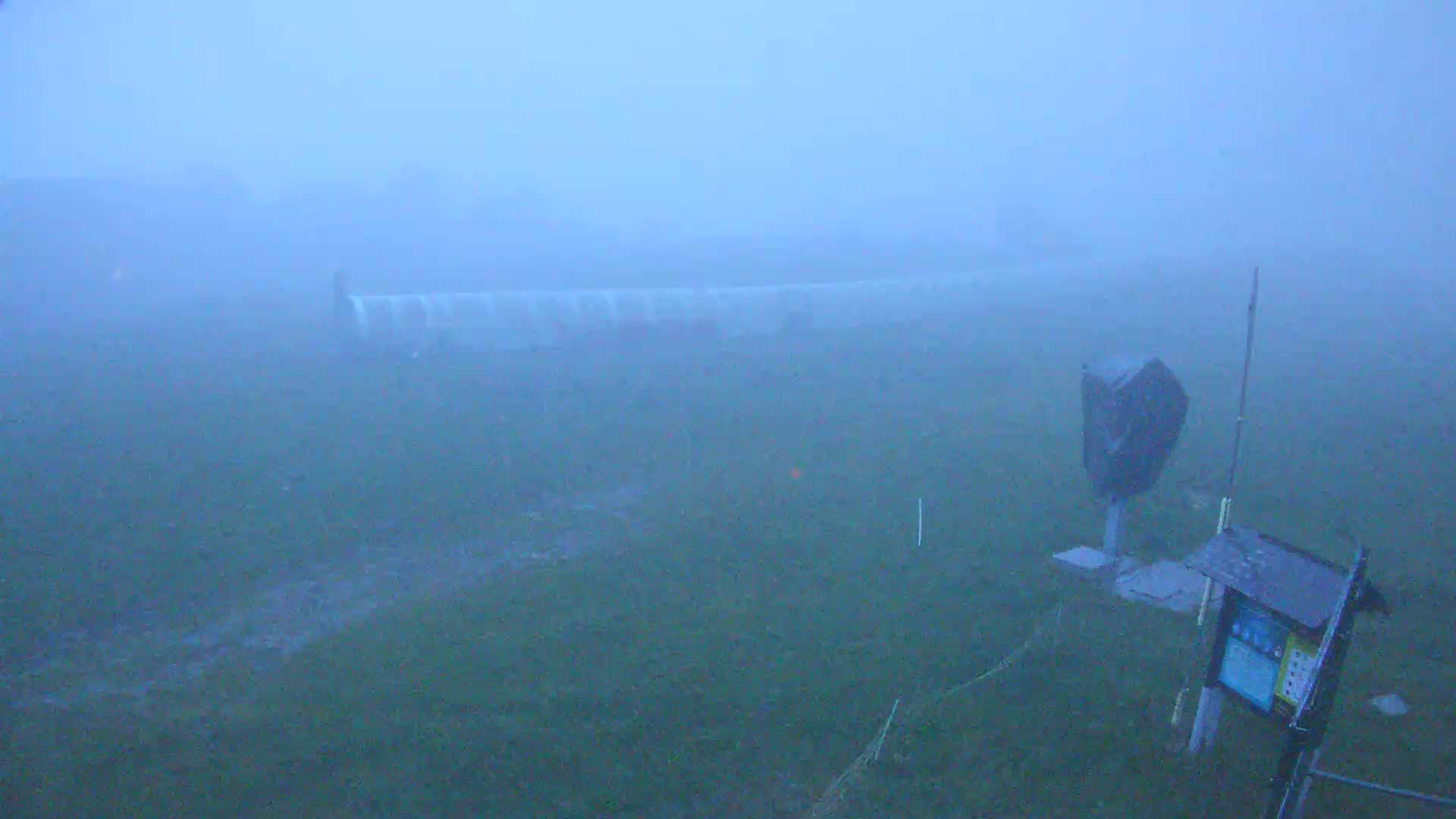 A foggy, outdoor scene shows a mostly obscured grassy field with a few pieces of equipment and a partially visible structure in the distance.