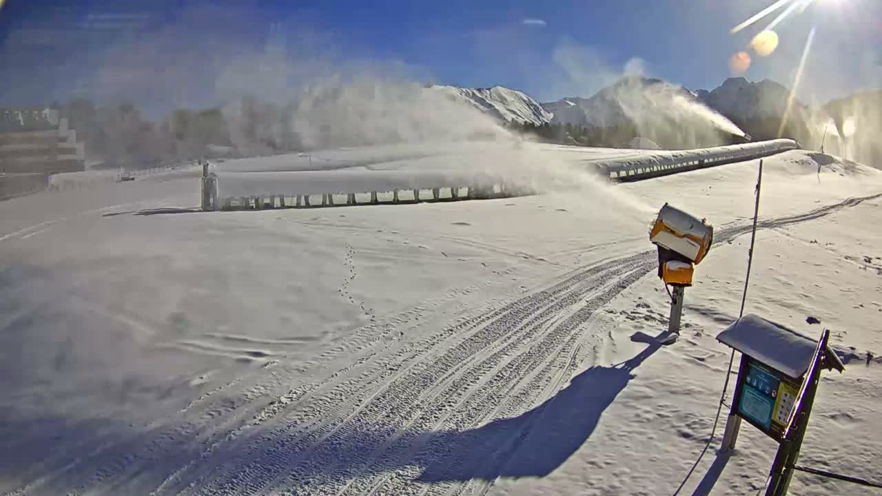 On a bright, sunny day with a clear blue sky, multiple snow cannons actively spray snow across a snow-covered mountain resort, featuring ski tracks, a long covered lift, and distant snow-capped peaks.