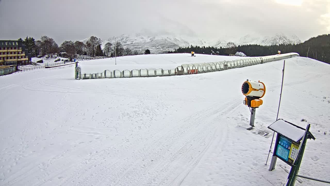 A wide, overcast view shows a snow-covered ski area featuring a long, clear-domed magic carpet lift, a hotel building, a snow cannon, and distant hazy mountains.