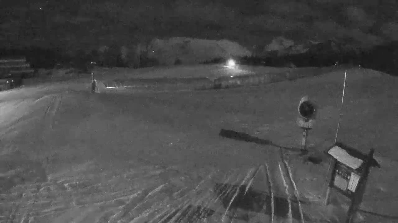 A snow-covered ski slope is visible at night, illuminated by distant lights, with tracks in the snow, a snow cannon, and a sign in the foreground under clear dark skies.
