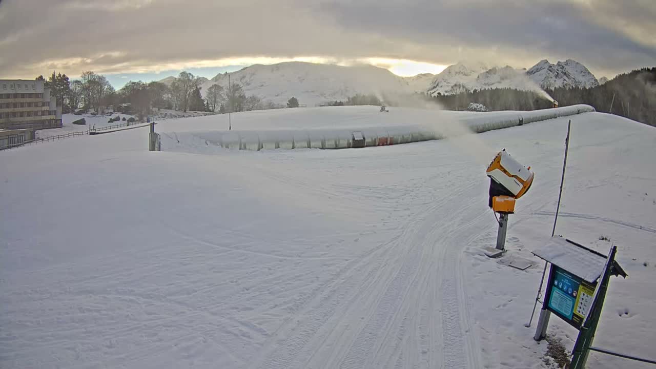 A snowy mountain ski area is seen under a cloudy sky with patches of brighter light, where multiple snow cannons are actively blowing fresh snow across the slopes with distant peaks visible in the background.