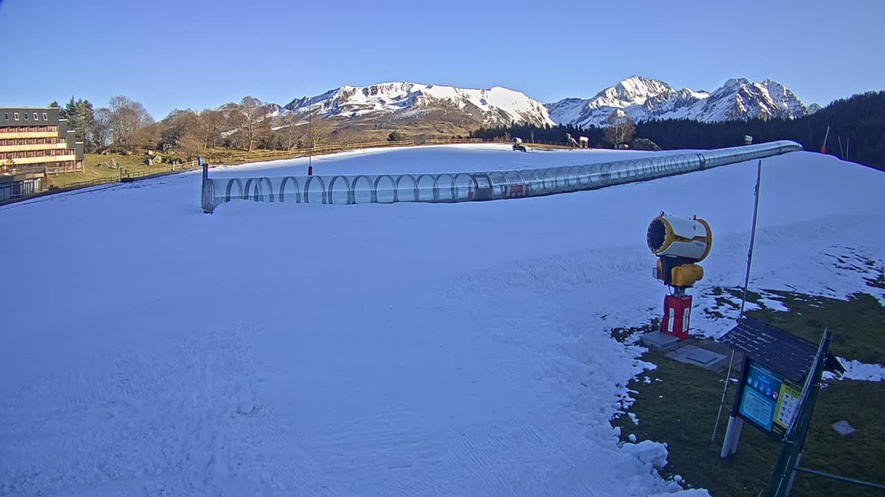 A snowy mountain ski area is seen under a cloudy sky with patches of brighter light, where multiple snow cannons are actively blowing fresh snow across the slopes with distant peaks visible in the background.