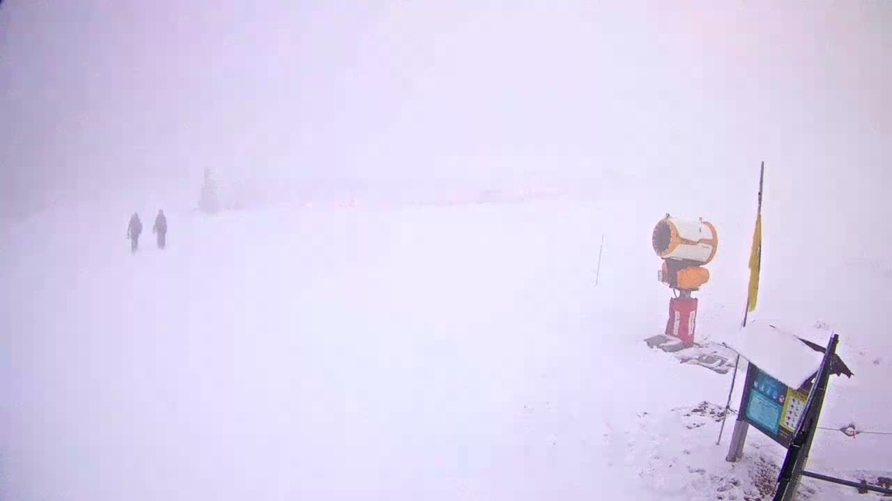 A snowy mountain ski area is seen under a cloudy sky with patches of brighter light, where multiple snow cannons are actively blowing fresh snow across the slopes with distant peaks visible in the background.