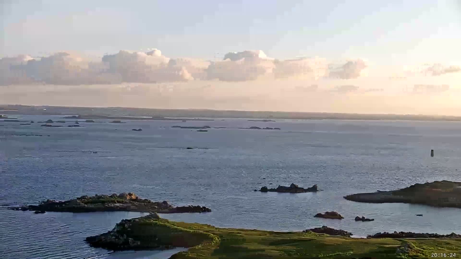 A tranquil coastal scene under a partly cloudy sky shows a wide expanse of water dotted with small rocky islands and a foreground of green land.