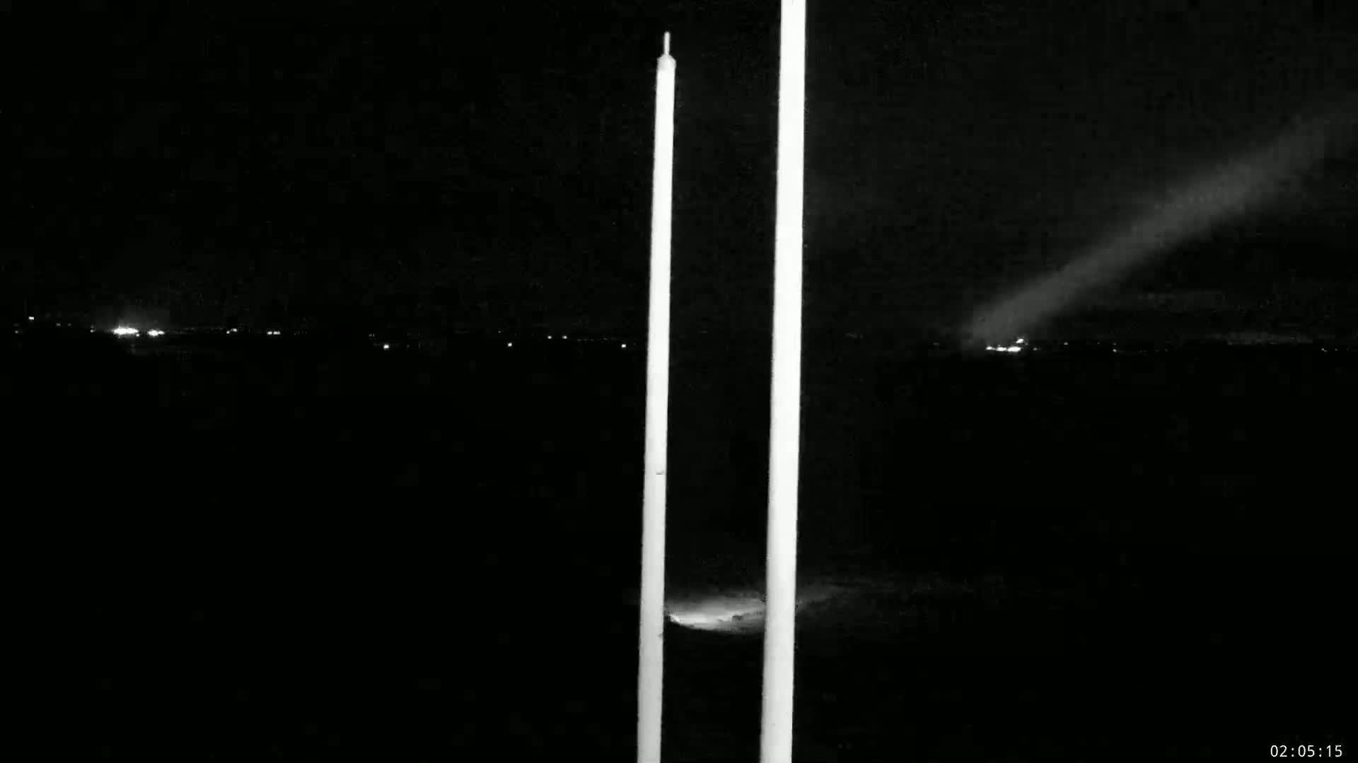 Two tall, thin, light-colored poles stand in the foreground of a nighttime scene with a bright light source visible in the distance, possibly a searchlight or distant city lights.