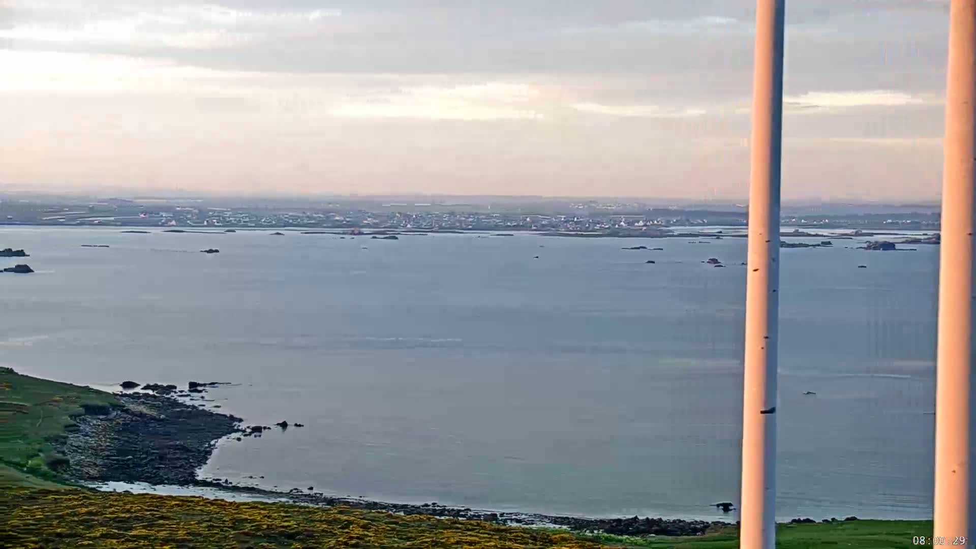 A calm, pale-blue sea stretches to a distant town under a soft, cloudy sky, viewed from a coastal hillside with two tall pale poles in the foreground.