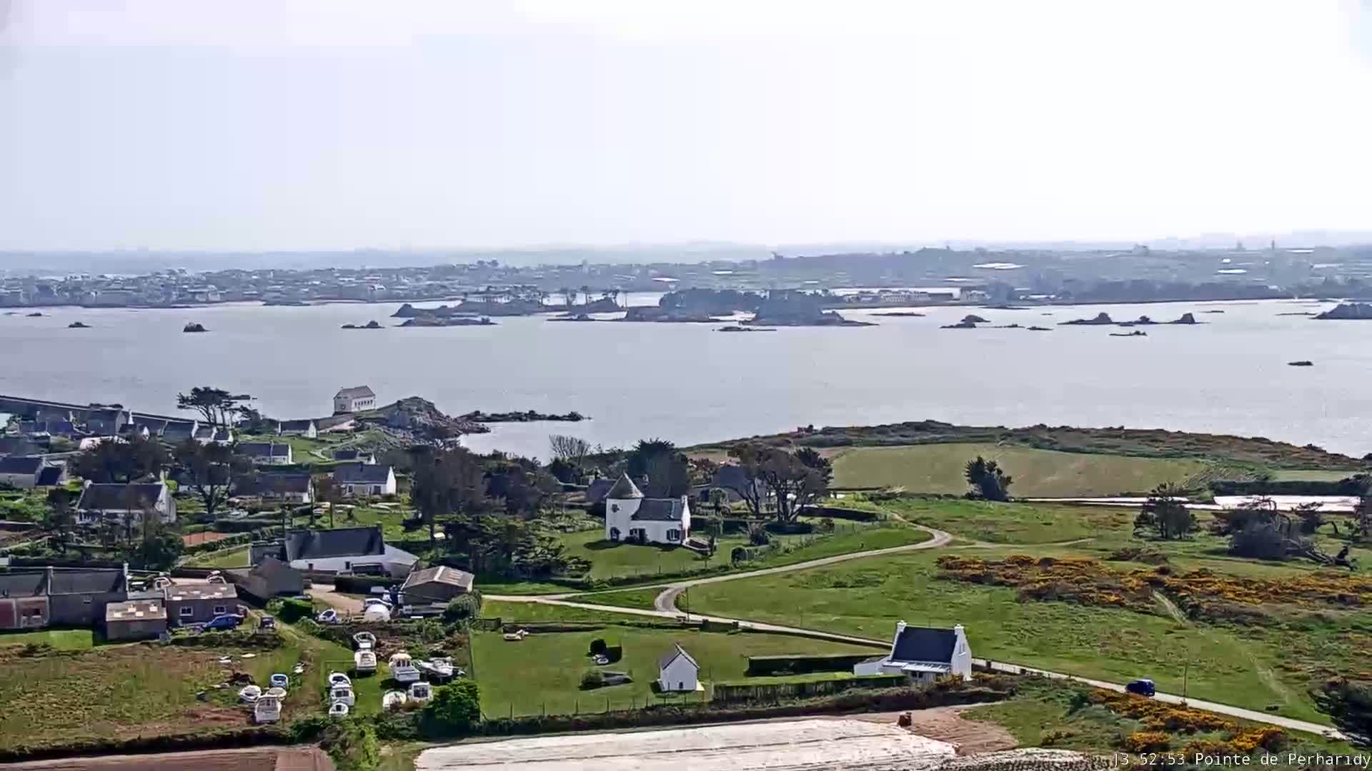 A coastal village with white houses and green fields overlooks a calm body of water dotted with small islands under a bright, hazy sky.