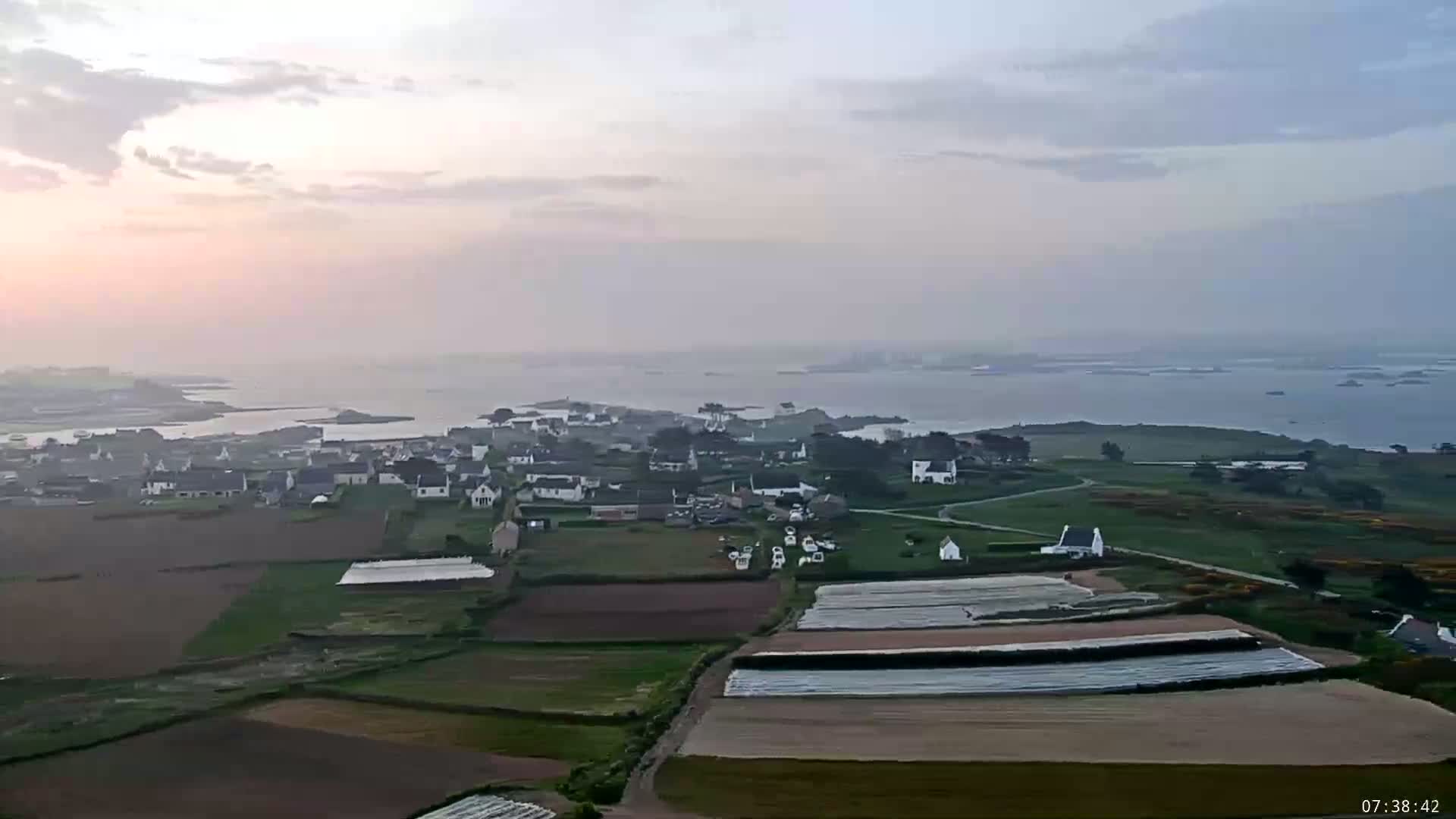 An aerial view shows a coastal village with surrounding farmland under a hazy, sunrise sky.
