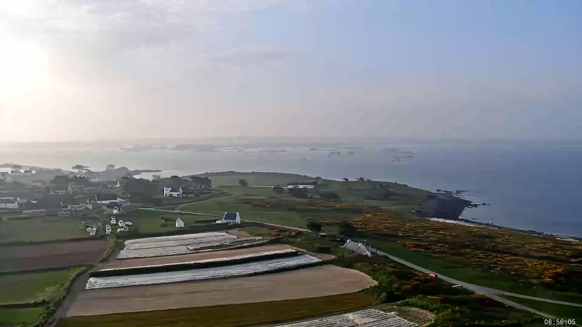 A hazy coastal village with farmland and a calm ocean under a partly cloudy sky.