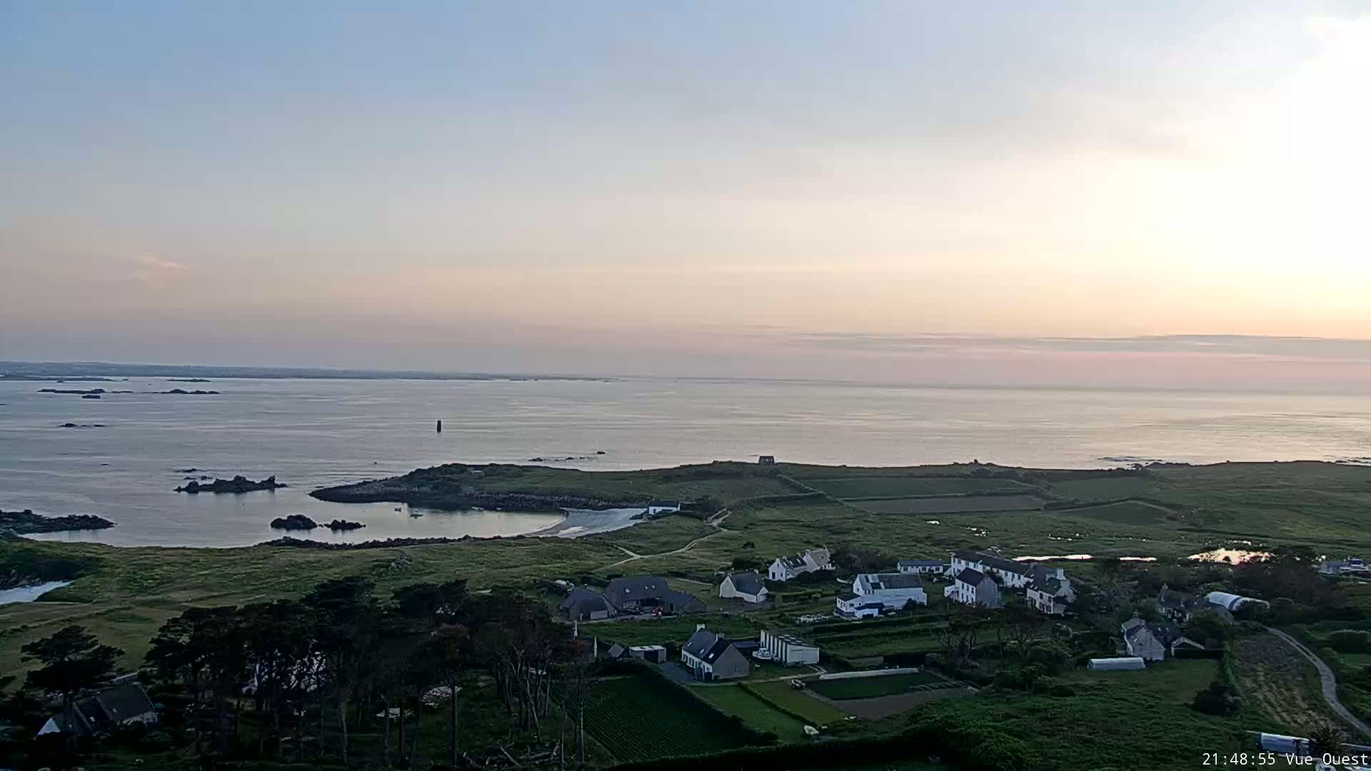A tranquil coastal village nestled on a verdant peninsula overlooks a calm ocean under a pale, sunset sky.