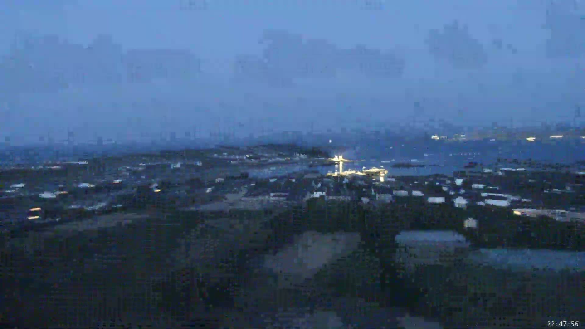 A coastal town at twilight is partially visible under a mostly cloudy sky, with a body of water in the background.